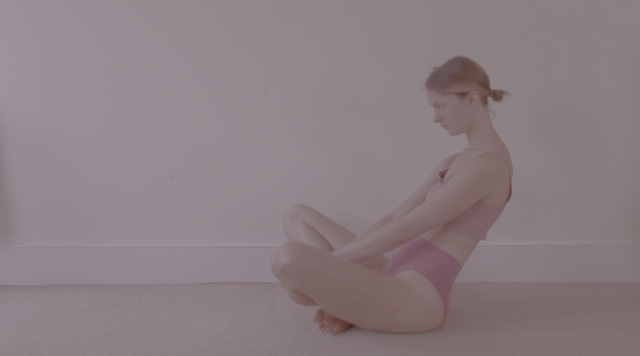 Susanna in profile, sitting on the floor with her legs crossed. She is rocking on her sit bones and leaning back, mid-movement.