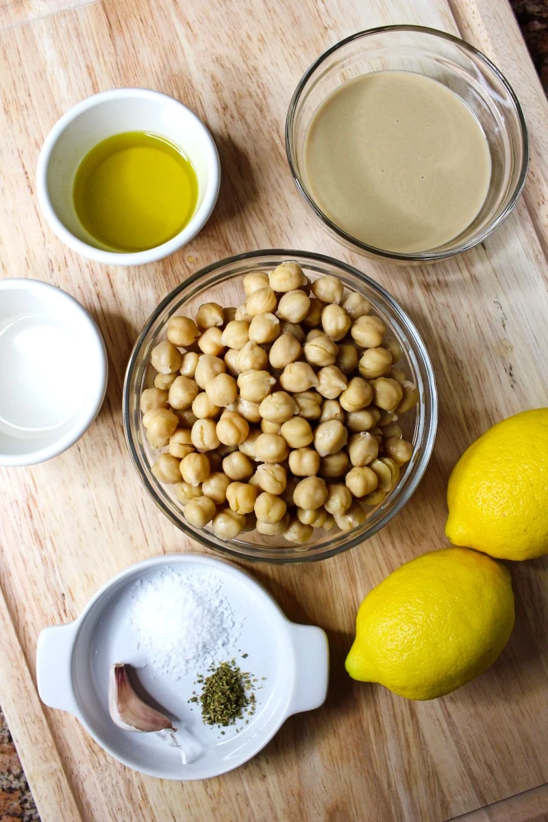 Small bowls of chickpeas, water, olive oil, and tahini, two lemons, salt, oregano, a clove of garlic, and two lemons arranged on a wood cutting board.
