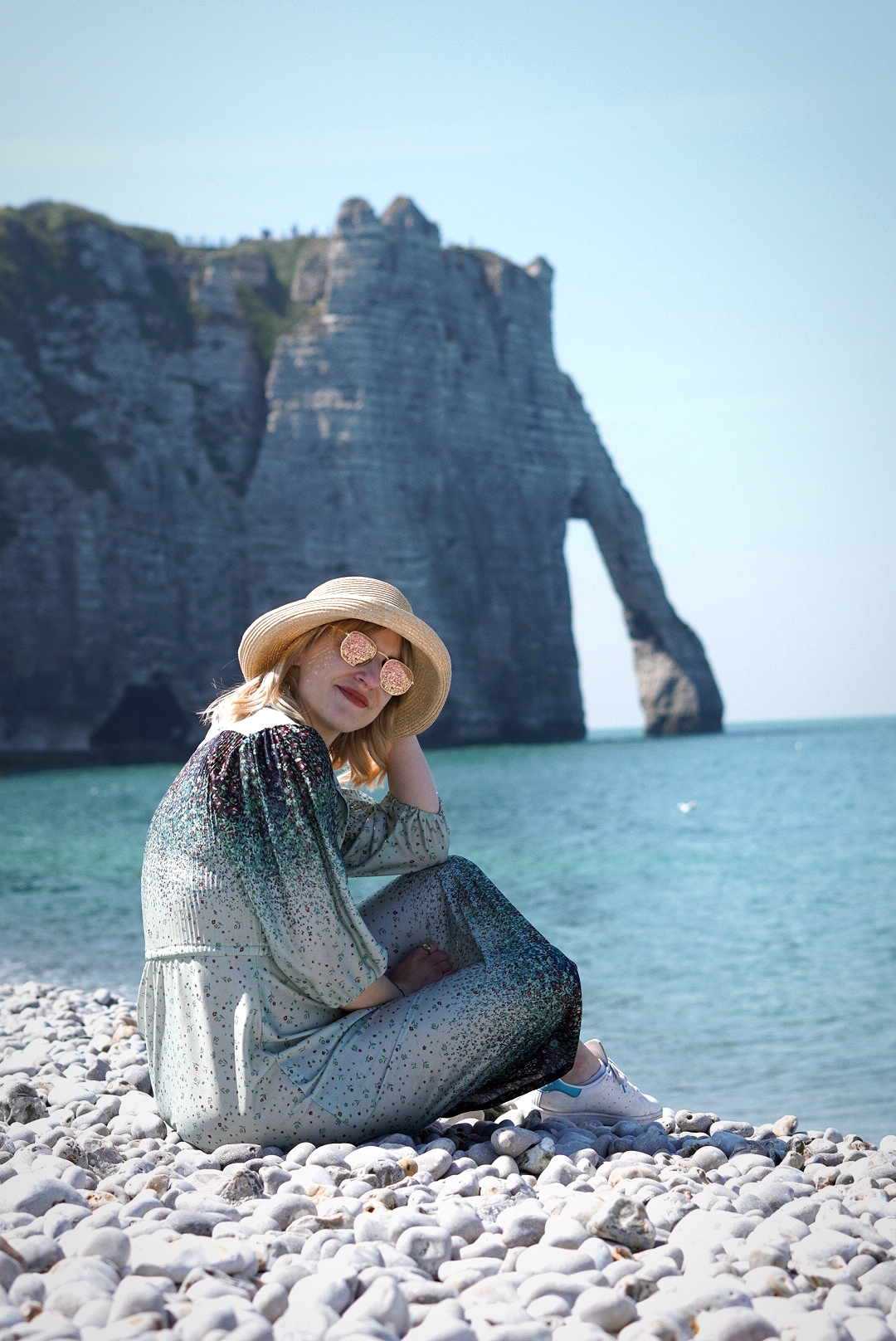 Amy is sitting on a pebbled beach looking back to camera, in the background is the sea and a huge cliffside. She is wearing a blue patterned dress and a straw sun hat