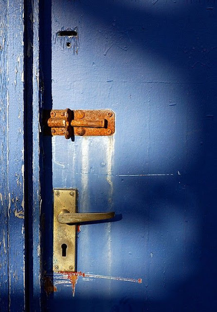 Part of a worn, chipped and scratched up wooden blue door with a gold handle at the bottom and a completely rusted up bolt above it. Slightly above that is a tiny opening from where there was another lock at some point. Taken by Steve  Hutch from CdL Creative