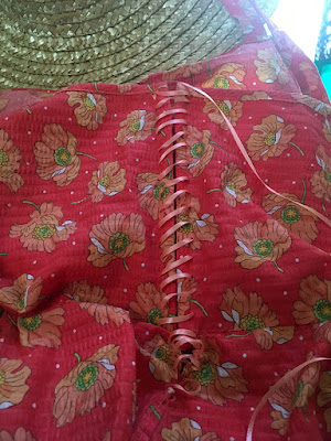 A close photo of the back of a coral sundress with large, scattered orange poppy motifs. Narrow peach satin ribbon laces the back edges together; at the top of the photo is the brim of a straw hat.