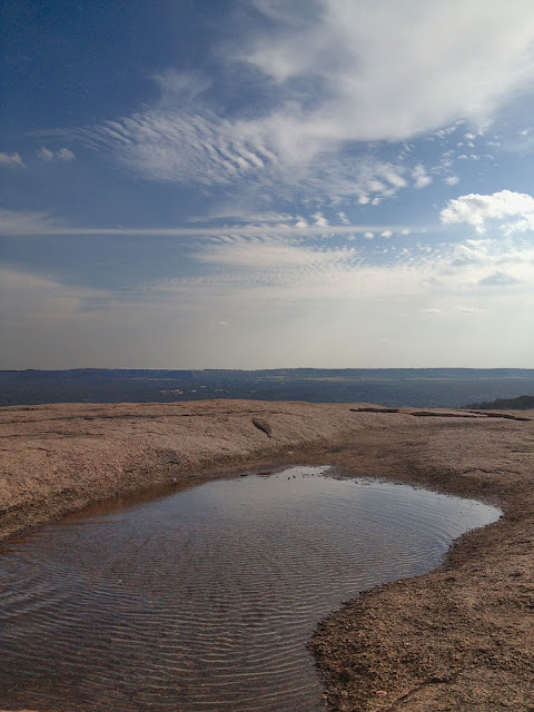 View of the Texas Hill Country from the top of Enchanted Rock. In the foreground of the photo a vernal pool ripples in the wind.