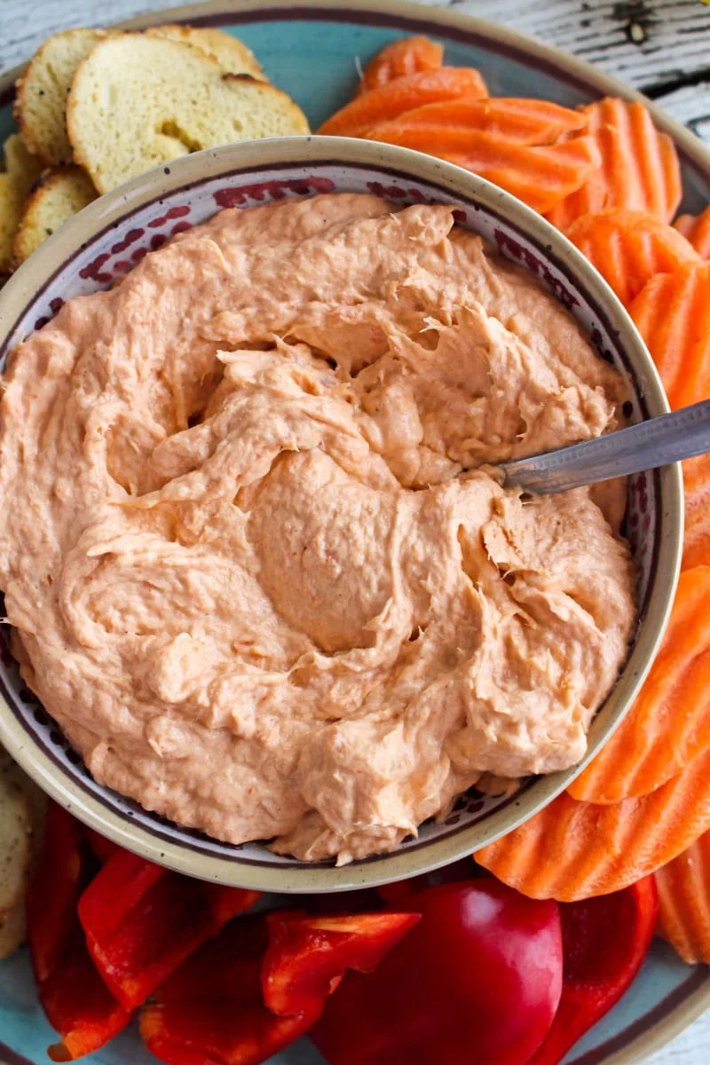 Closeup top view of Smoked Salmon Dip in a tan bowl on a blue plate with bagel chips, carrot chips, and bell pepper pieces around it.