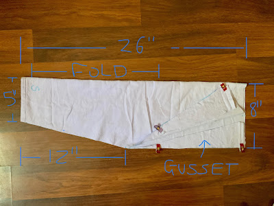 A sleeve piece cut out of lavender coloured fabric is laid out on a wooden floor, folded in half lengthwise. The photo has been marked up with measurements. The sleeve is 26" long in total, and 8" wide at the fold. Triangular pieces have been cut out of the sleeve to make it taper from the elbow to the wrist. The wrist end is 5" wide at the fold and the tapered cut is 12" long. The upper part of the sleeve has been folded back at the cut sides to show the triangles that were cut off the sleeve taper have been rotated and pinned to create an extra triangle of fabric at the upper sleeve edge. The diagonal edges of each triangle have been placed along the straight edge of the sleeve pieces. End ID.