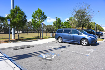 A blue minivan is backed into an accessible parking spot. There is another spot, which is empty, located closer to the camera and on the opposite side of the unloading zone. There is a sidewalk that runs behind the minivan, a series of trees behind it, and then a fenced in field that's located behind that.