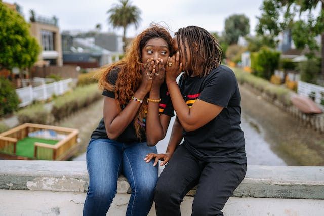 Two young women sitting on a bridge whispering to each other