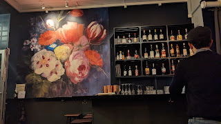a man sits at a bar, behind the bar are neatly organised bottles and to the left a huge floral mural on the wall