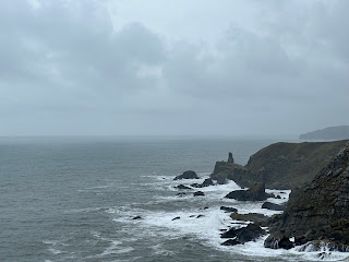 A view of a grey looking rocky coastline with ominous clouds above and waves crashing against the cliffs. Photo by Kevin Nosferatu for the Skulferatu Project.