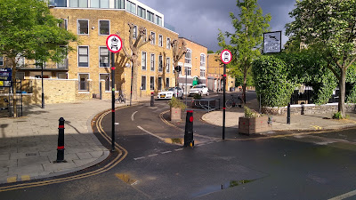 A view of the filter which has trees to the left, bollards and traffic signs stopping motor traffic and planters to the right.