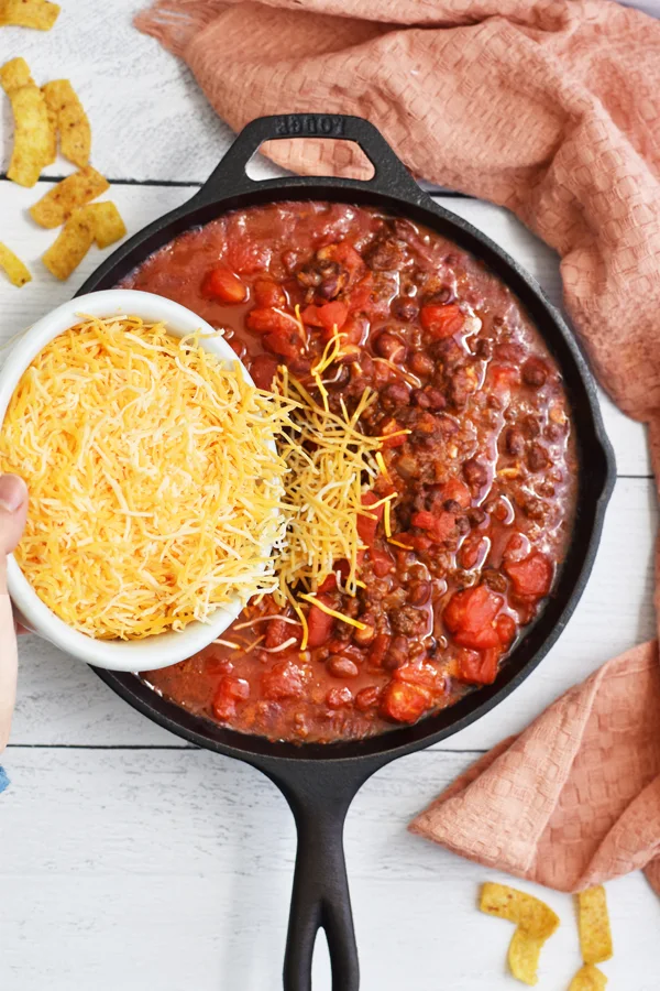 Shredded cheese being sprinkled from a white bowl onto a cooked beef mixture in a cast iron skillet, melting and creating a delicious cheesy topping.