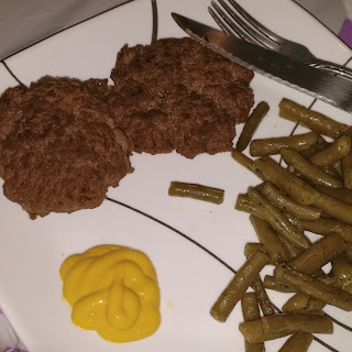 Two hamburgers on a plate with sautéed green beans. A dollop of mustard is on the plate alongside the hamburgers. A knife and fork are at the upper right corner.