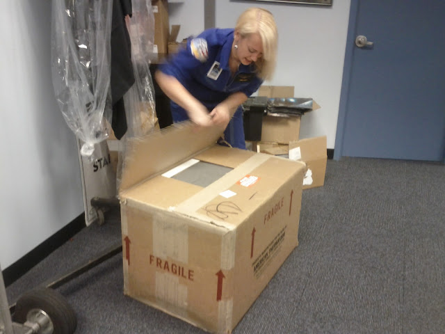 A 5'4" blonde woman in a blue flight suit is opening a very large brown cardboard package. The package is about three feet tall and two feet wide and across.