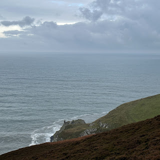 A photo looking down a hill towards the sea.  In the distance can be seen a piece of land jutting out with some ruins on it. Photo by Kevin Nosferatu for the Skulferatu Project.