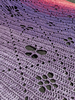 An angled photo of the crochet shawl flat on a couch cushion; the shawl is gradient from pale purples, to darker, and changes to pink towards the top. There is a design created by holes of a grid with motifs of butterlies, flowers and dragonflies within.