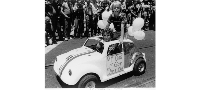 Two young boys in a toy VW Beetle with a sign reading "My Dad is Gay and That's Okay" in SF pride parade ca. 1977