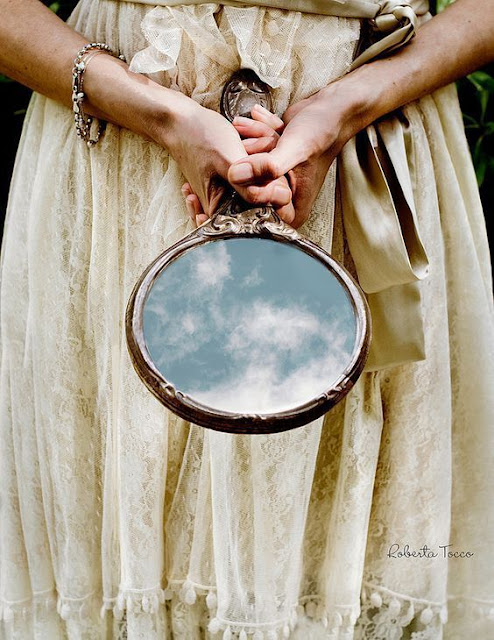 A woman in a cream dress holding a round handmirror that is reflecting the sky - taken by Roberta Tocco