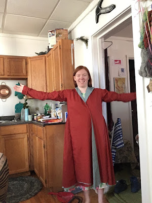 A young redheaded white woman, standing in the doorway between a messy kitchen and bedroom, and wearing a long-sleeved red caftan that comes just past her knees, with a grey-blue lining showing at the folded-over neckline and front overlap.