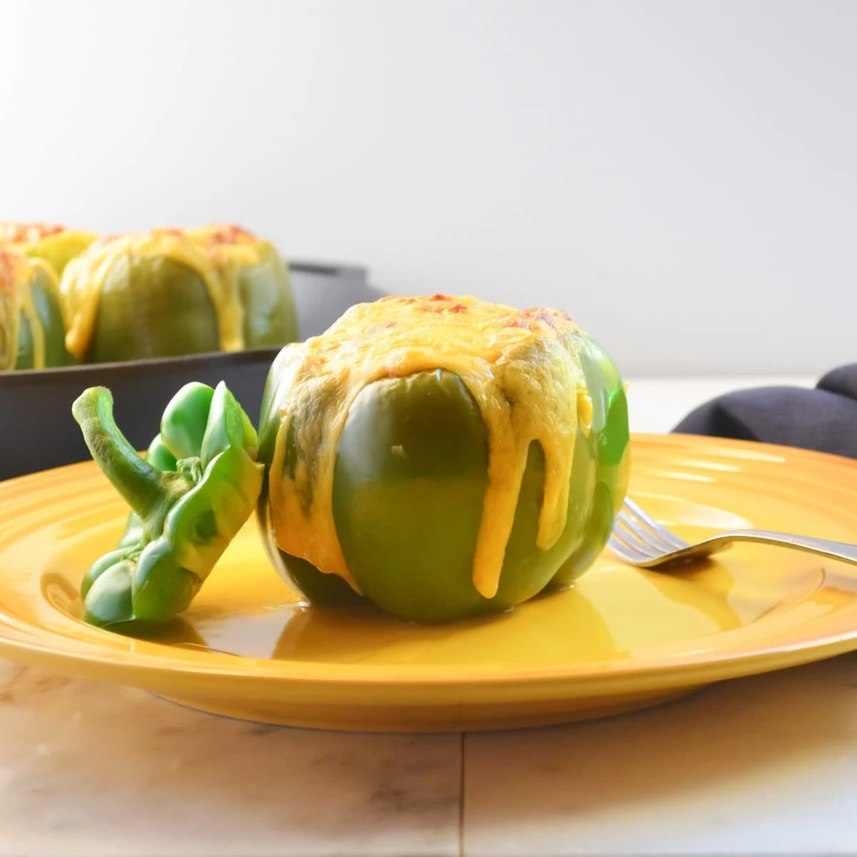 A side shot of a Stuffed Bell Pepper on a yellow plate with a cast iron skillet full of Stuffed Bell Peppers on a white marble counter top.