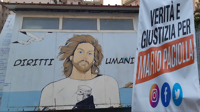 Sullo sfondo il murale di "Humanhero Samuel" non lontano da dove viveva Mario, a Napoli. A destra lo striscione del gruppo "Giustizia per Mario Paciolla"