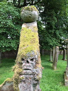 A photo of a sinister looking skull carved on the side of a graveyard.  It is lichen covered and the stone rises up from its head like an old fashioned hat.  Photograph taken by Kevin Nosferatu for the Skulferatu Project.