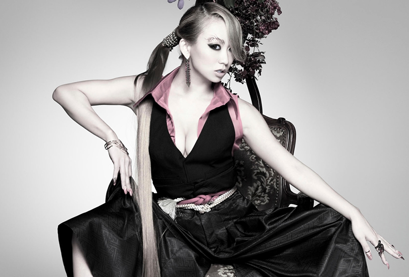 Koda Kumi is sat on a chair, wearing a black sleeveless suit outfit, with flared trousers and a pink sleeve shirt underneath. Her hair is in a long ponytail, which runs down in front of her on her right-hand side.