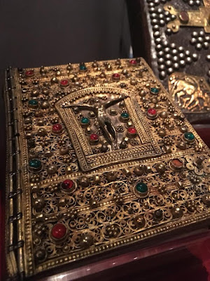 A gold book cover, completely covered in dense spiral filigree, raised gold beads, and inset polished carmine and emerald gems. In the center, an arched rectangular frame contains a raised gold figure of Christ on the cross, also surrounded by raised gold beads and inset carmine and emerald gems.