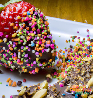 Image of a chocolate covered strawberry being sprinkled with rainbow sprinkles and chopped nuts.