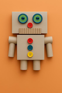 A cute robot figure made from recycled cardboard materials is displayed on an orange background. The robot has a rectangular head and body, button eyes and controls, a corrugated cardboard mouth, and arms and legs made from paper tubes, showcasing a creative and eco-friendly kids’ craft.