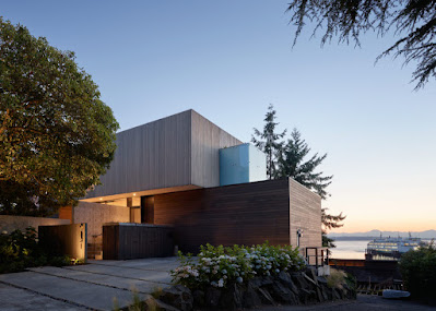 Image of Fauntleroy Residence from the street. Showing driveway and wood-covered garage portion, and concrete walls leading to front door. Upper floor is an offset box with vertical wood siding. A small opaque glass box is attached to the upper floor, above the garage. A large deciduous tree is in the front year; portions of coniferous trees appear behind the house. In the distance is the view of the bay and the Southworth - Fauntleroy ferry terminal