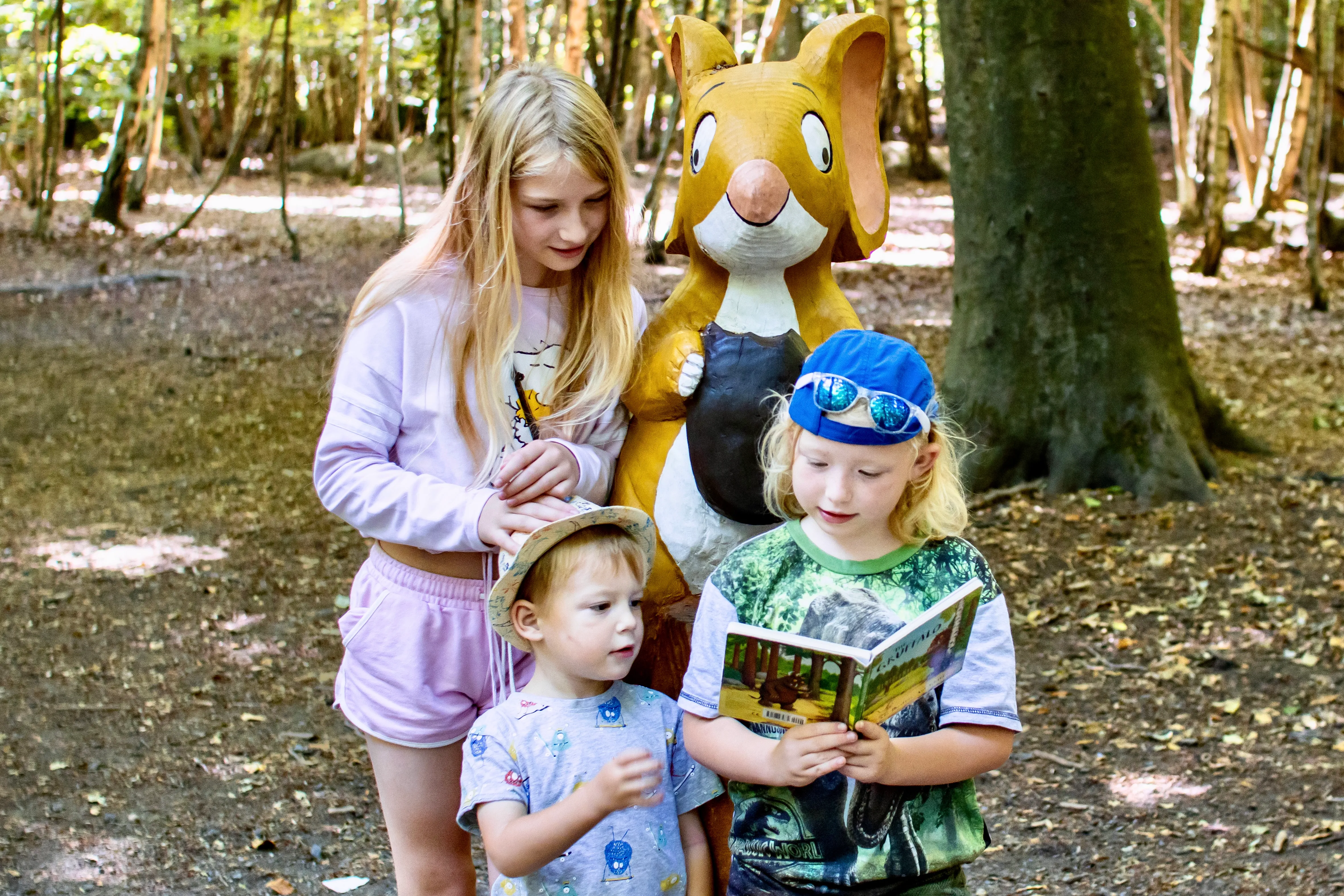 3 children standing next to a wooden carving of the mouse from the gruffalo reading the gruffalo book by Julia Donaldson