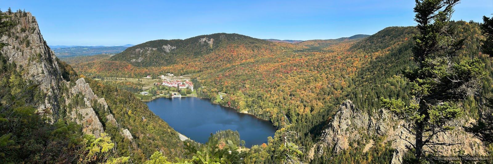 Dixville Notch landscape photograph containing jagged rock cliffs, green trees and the beginning of fall colors. The Balsams Resort is visible behind Lake Gloriette.