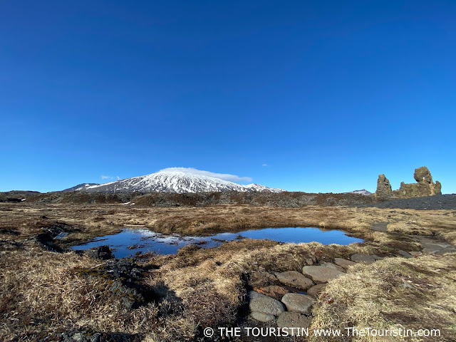 A snow-capped volcano partly reflected in a small pond, and two large basalt cliffs surrounded by rocky terrain under a bright blue cloudless.