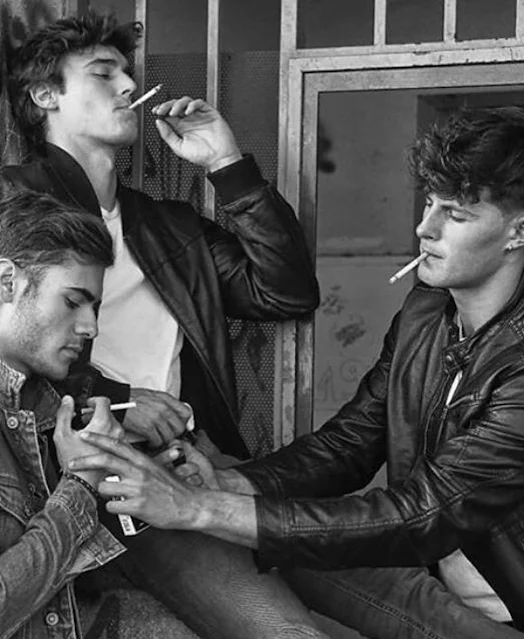 Black and white photograph of three guys smoking cigarettes wearing black leather biker jackets one dude is lighting the other cigarette