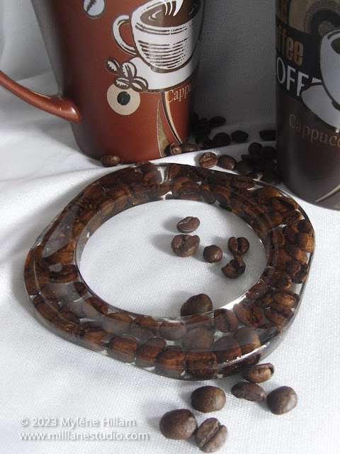 Square resin bangle bracelet filled with coffee beans, sitting on a white table top, in front of two coffee mugs