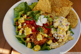 image of a bowl of taco rice topped with shredded cheese, diced tomatoes, corn, lettuce, and tortilla chips
