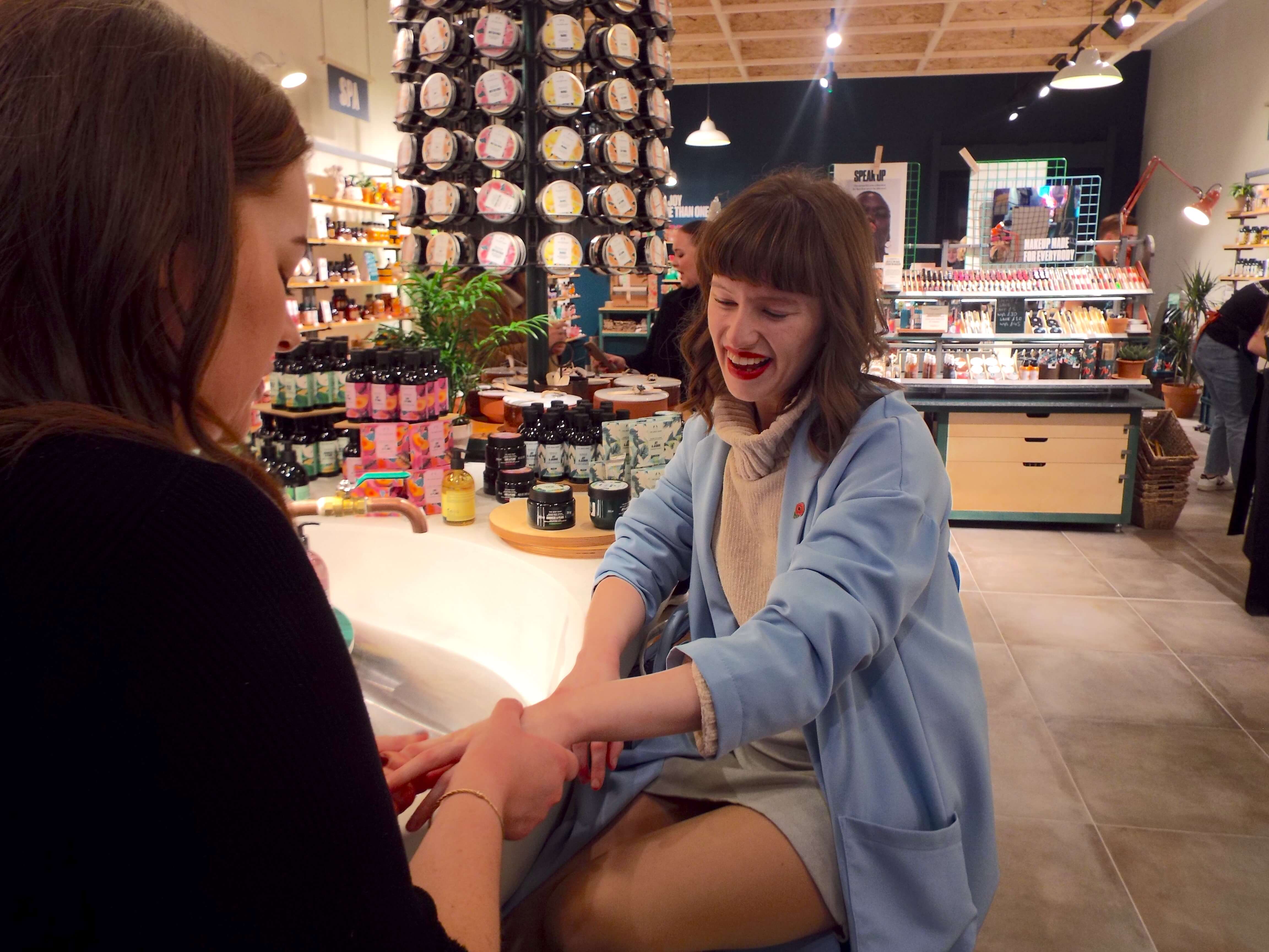 Ellie sat at the product testing island, getting a hand massage and pamper, smiling and talking with Lauren, the beauty assistant performing the hand massage.