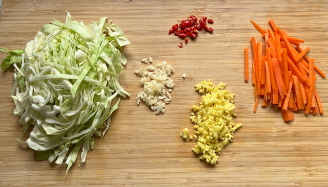 A cutting board with sliced cabbage, matchstick carrots, slices chili peppers, minced garlic and ginger