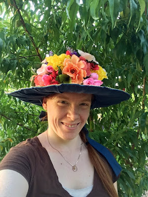 A selfie of a redheaded white woman smiling into the camera. She's wearing a brown v-neck t-shirt with a white camisole, and a thin gold necklace with a white gemstone pendant and thin gold hoop. Her hair is braided with a long tail that falls over her left shoulder, and she's wearing a wide-brimmed hat lined in medium-blue fabric, and piled so heavily with bright-yellow, peach, pale-pink, coral, and purple flowers that the crown is completely hidden. A long medium-blue fabric tie falls over her hair, and the background is filled with branches carrying glossy green, pointed oval leaves.