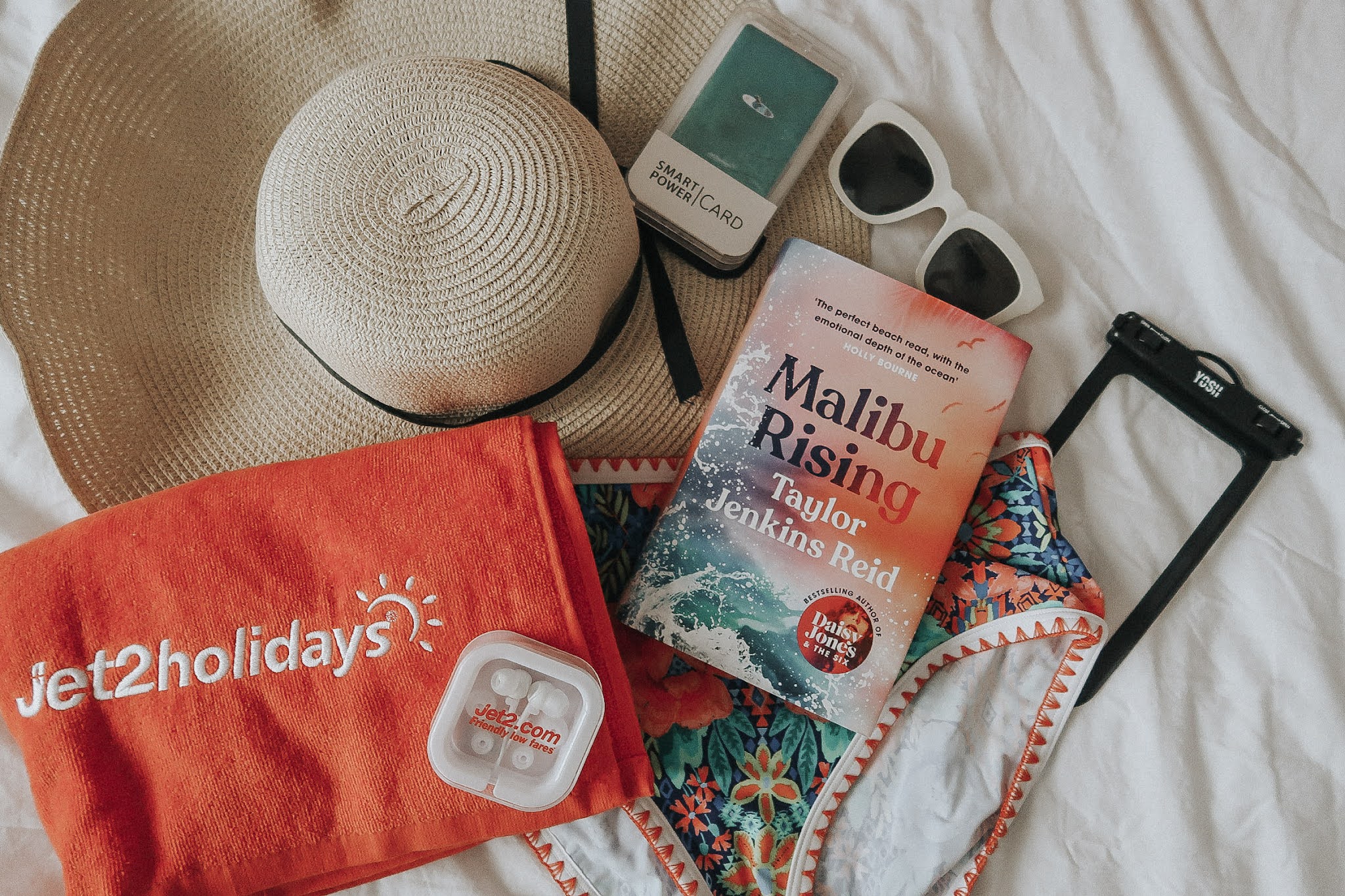 A red beach towel, a pink book, white sunglasses and large beach hat.