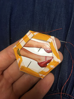 A closeup of a paper hexagon showing tan legs in shiny red high heels. Yellow fabric with irregular white stripes wraps around from the other side of the paper, covering a narrow border aorund the shape. Red thread shows in small basting stitches at each corner, and long runs down each side; the needle, still threaded, is just visible in the background on the photographer's denim skirt.