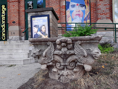 The top of a column sitting on a lawn next to a walkway. It is decorated with fierce looking animal heads.