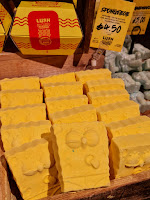 A large rectangular light brown box with a row of yellow square pantless bath bombs with animated eyes and a sausage nose engraved with a smile into the bomb with lush engraved into the bottom next to a small rectangular yellow label card with soongebob bath bomb lush in black font on a bright background