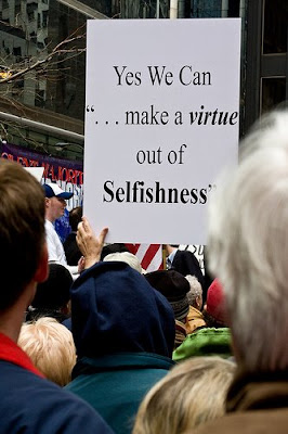 protester with sign saying yes we can make a virtue out of selfishness