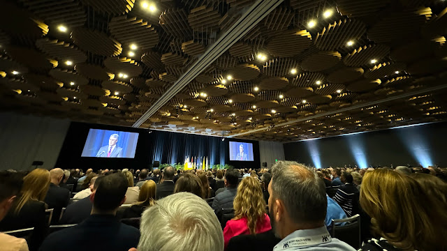 A large ballroom full of people, a man stands on stage and two large screens are on either side of him