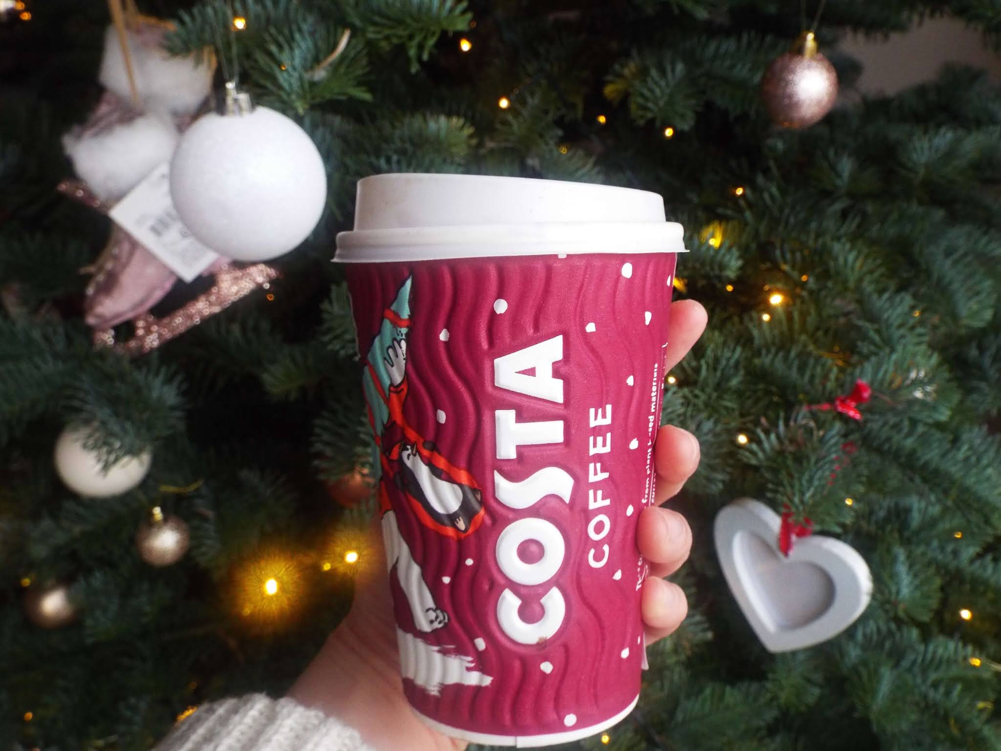Secret recipe vegan Costa Christmas drink, displayed in Costa Christmas Cup with polar bear carrying Christmas tree. Held up in front of cosy, warm white light Christmas tree, with an arm extending wearing a cream chunky knit jumper.