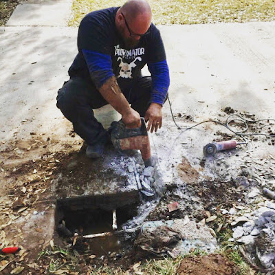 A plumber holds a concrete drilling tool, making a square hole in the driveway bigger. The hole is about 2 feet by 2 feet and shows exposed water pipes