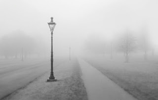 A grey photographic image of a lamp-post at the side of a path between areas of grass leading away from the frame. There are some faint outlines of trees in the foggy middle-distance.