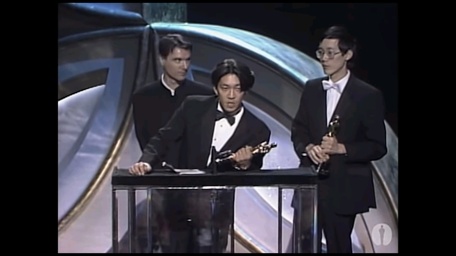 アカデミー賞の作曲賞の受賞式でスピーチを行う坂本龍一とその後ろに立つ共同受賞のデヴィッド・バーンと蘇聡
