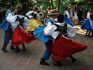 A photo of folkdancers wearing traditional clothes and dancing on paving stones under trees. The women are wearing colorful skirts in red, yellow, or blue; black bodices with colorful embroidery; and white blouses with very full sleeves. The men are wearing blue trousers, long blue vests, full-sleeved white shirts, and narrow-brimmed straw hats.