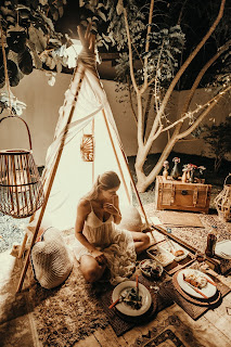 A sepia-toned picture of a woman sitting on the ground in a small teepee with plates of food on the rug around her.